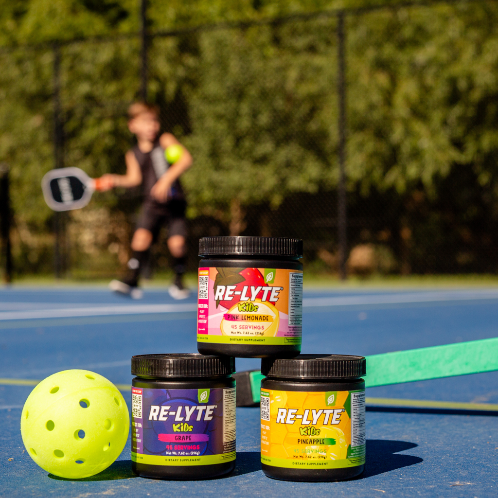 Three tubs of Re-Lyte Kids electrolyte powder in grape, pink lemonade, and pineapple flavors sit on a blue pickleball court with a pickleball and paddle in the foreground and a child playing pickleball in the background.