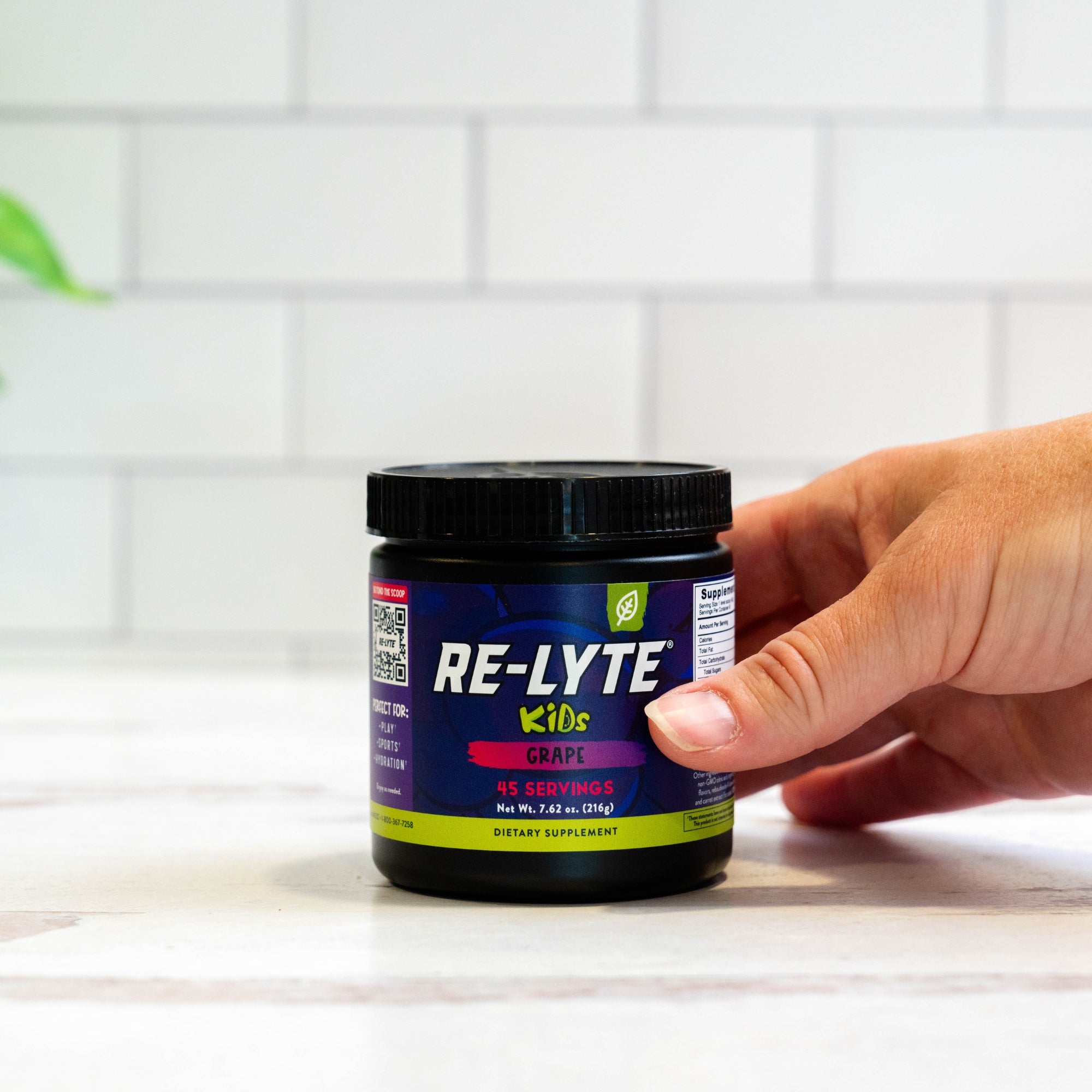Hand holds a black jar of Re-Lyte Kids Grape dietary supplement on a white countertop. 
