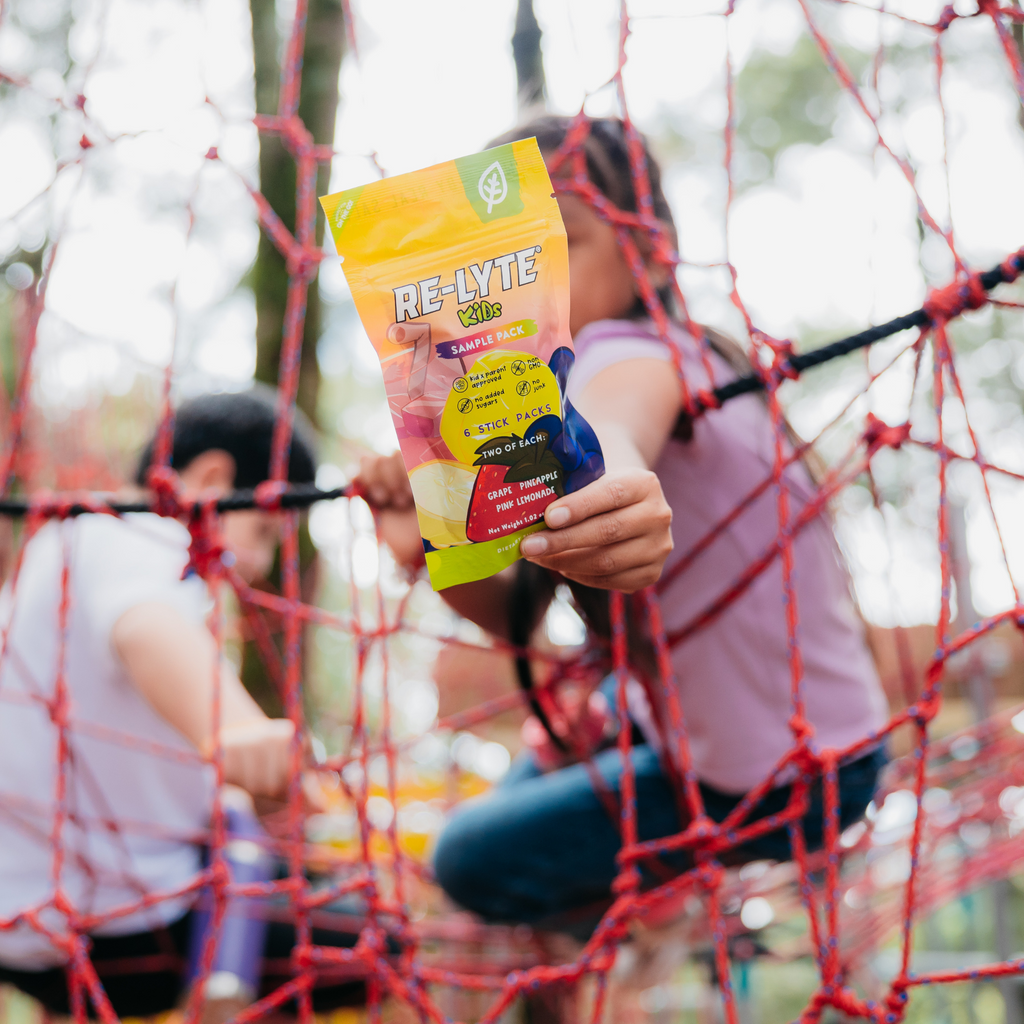 Child holds out a bright yellow sample pack of Re-Lyte Kids electrolytes against a blurred background of playground equipment.